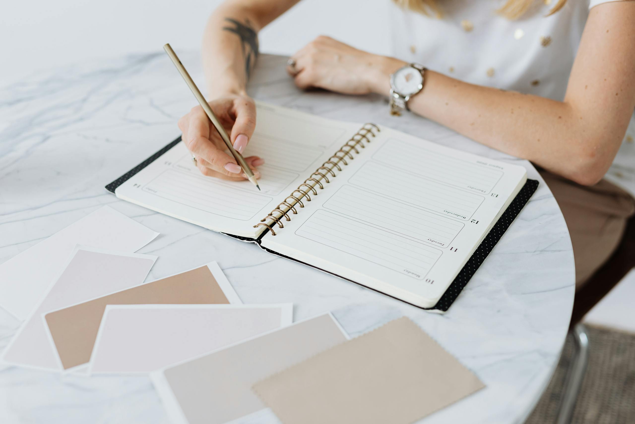 A woman writes in a planner on a marble table, surrounded by swatches. Creative and organized workspace.