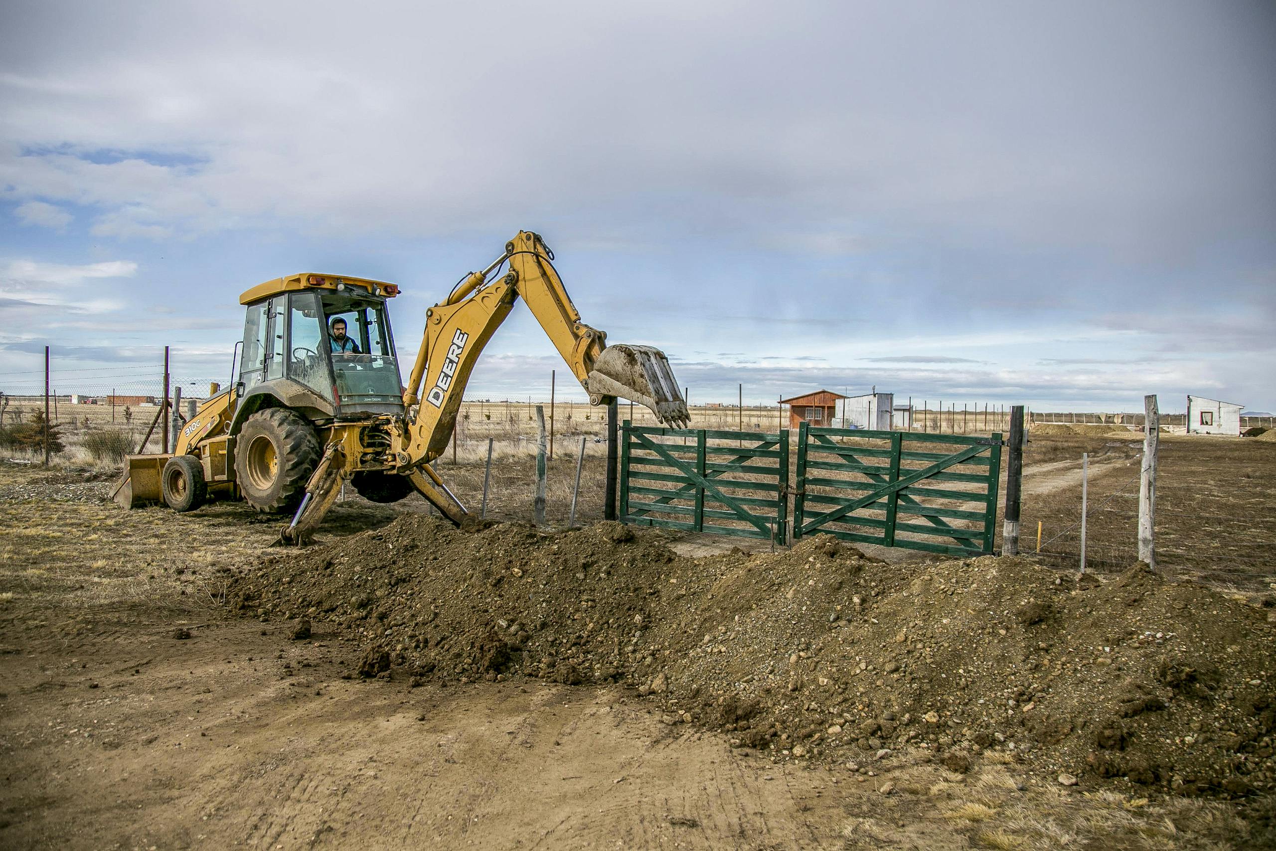 A yellow backhoe loader actively digs soil at a farm entrance with a green gate, under a cloudy sky.